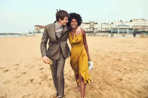 Man in brown suit with arm around shoulders of lady in yellow dress walking along beach. In the background round beach cafe and seafront buildings